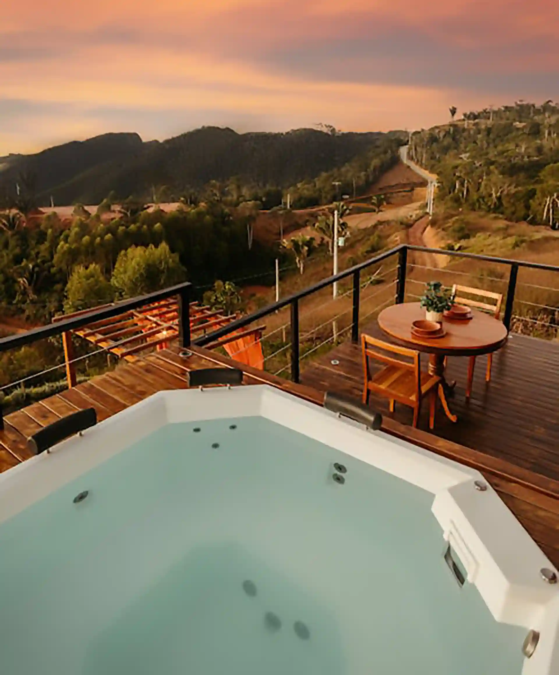 A hot tub on a wooden deck with a table and chairs, overlooking a scenic view of mountains during sunset.