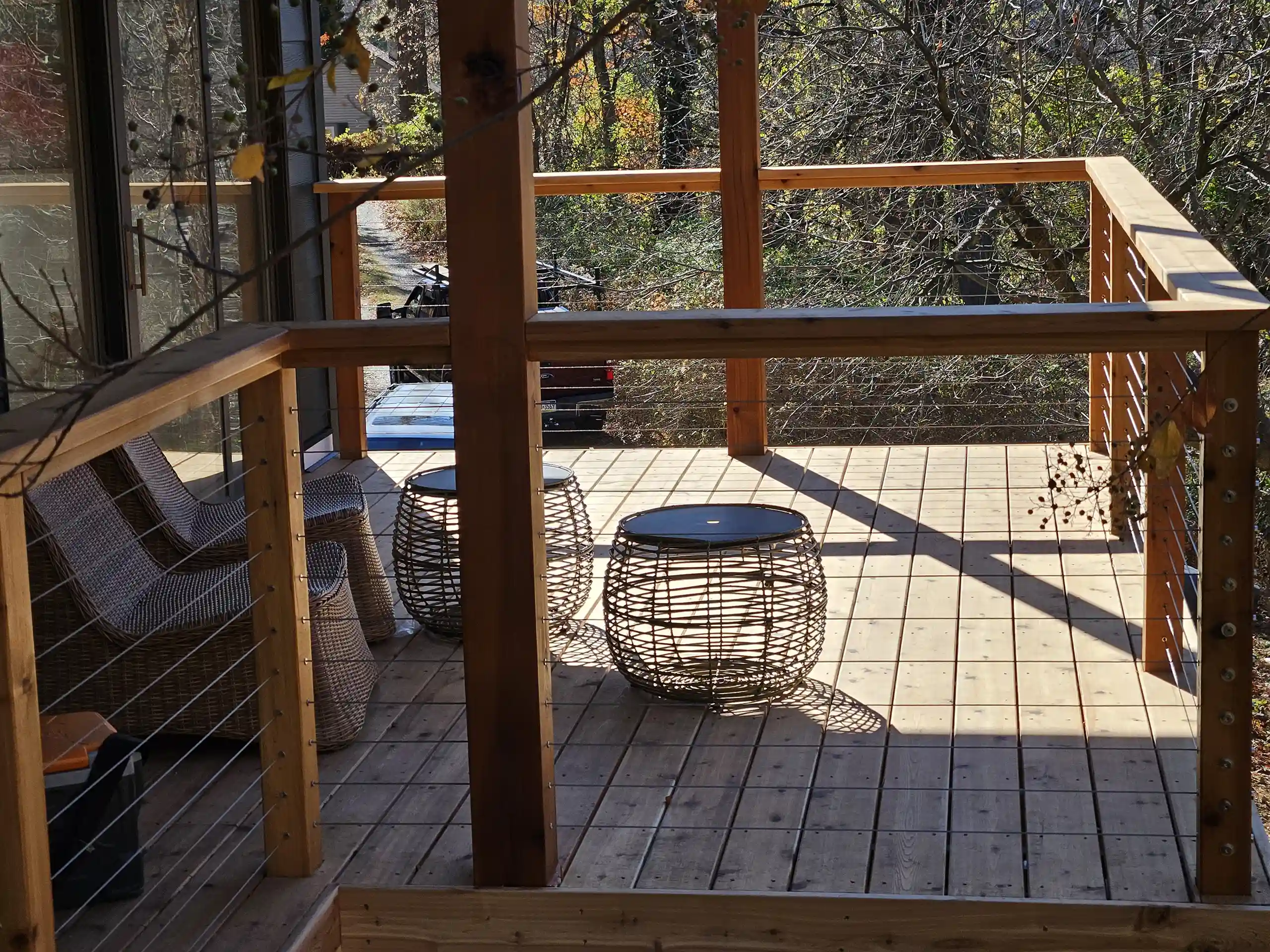 A wooden deck area with cable railing, two wicker chairs, and two round wicker side tables. Trees and a vehicle are visible beyond the deck.