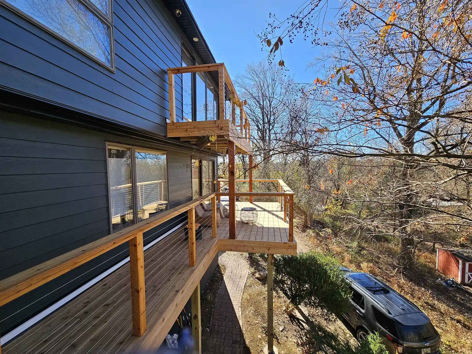 A multi-level wooden deck attached to the side of a house, with clear glass railings and vertical wooden posts. 