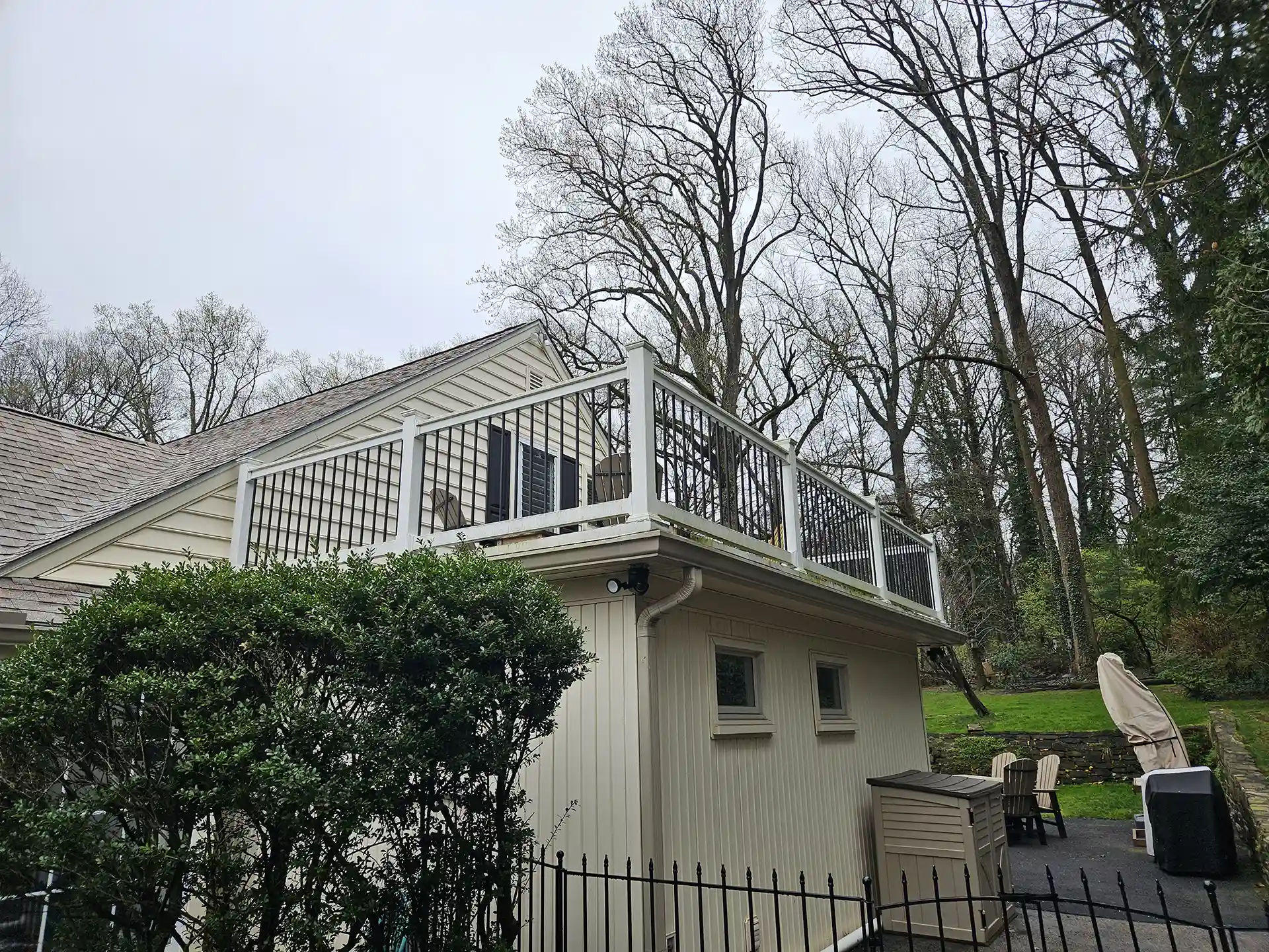 A second-story deck with black metal railings attached to a light-colored house. The deck overlooks a backyard with trees, shrubs, and outdoor furniture. 