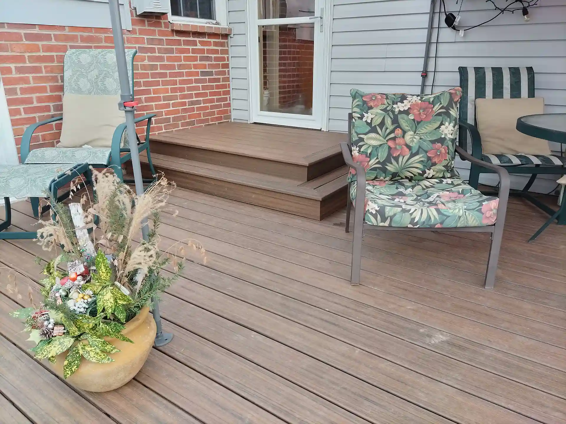 Composite deck with wood-grain texture featuring steps leading to a door, outdoor chairs with floral and striped cushions, and a planter with greenery. 