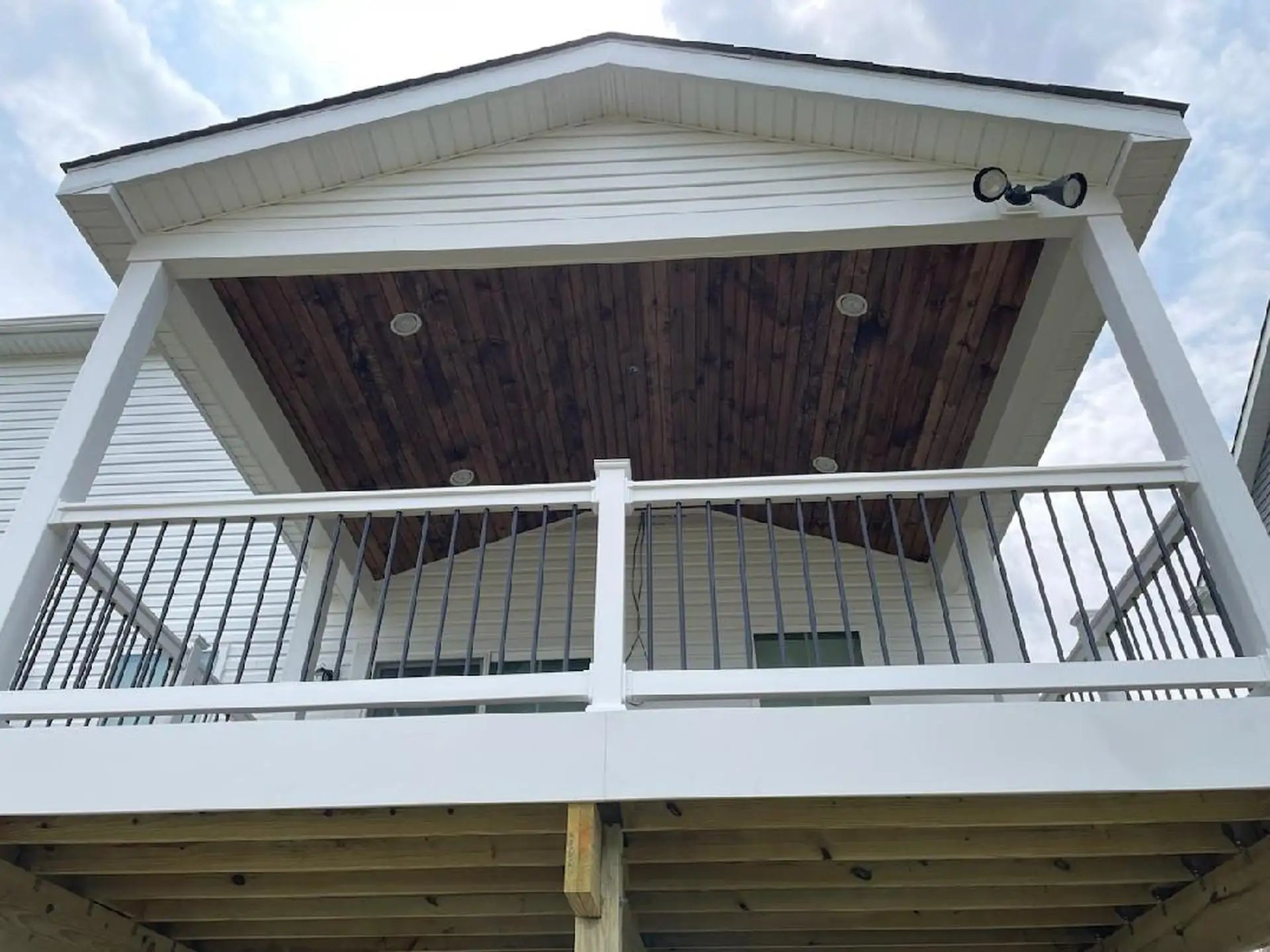 View of a covered deck with a wooden ceiling, white railing, and black metal posts. The space features recessed lighting and is elevated above the ground.