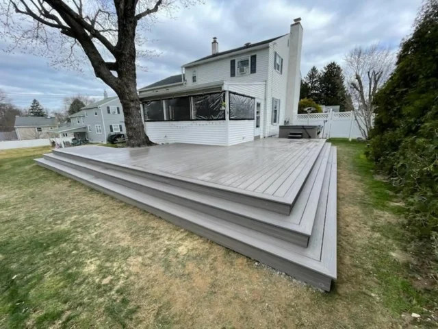 5 Reasons to Build a Multi-Level Deck for Outdoor Space A wide, ground-level composite deck with three-tiered, full-width steps. The deck boards are a light gray, and the stair risers are a darker gray. The deck is attached to a two-story white house with a large tree next to it. A hot tub is visible in the corner of the deck.