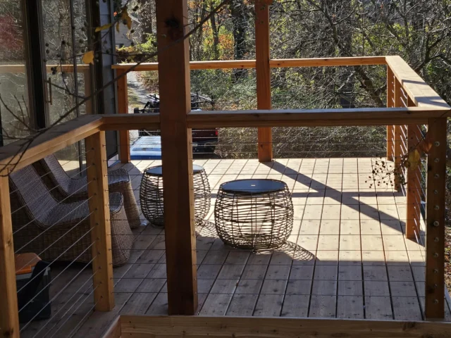 An outdoor deck with wooden flooring and railings. On the deck, there are two woven chairs and two woven, cylindrical tables. The deck appears to be surrounded by trees.